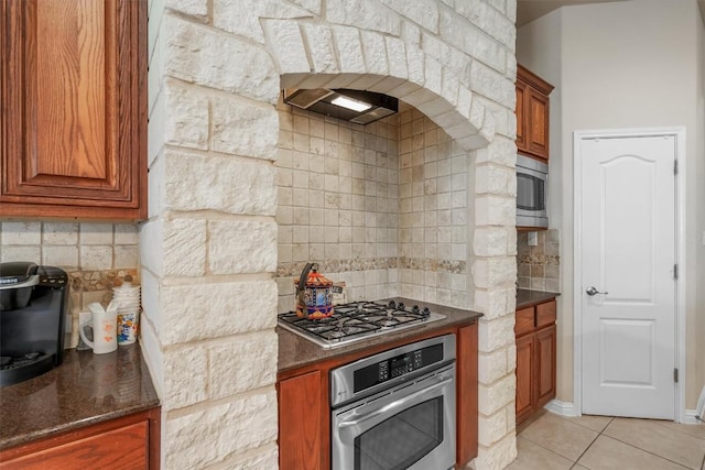 kitchen featuring backsplash, light tile patterned flooring, dark stone counters, and appliances with stainless steel finishes