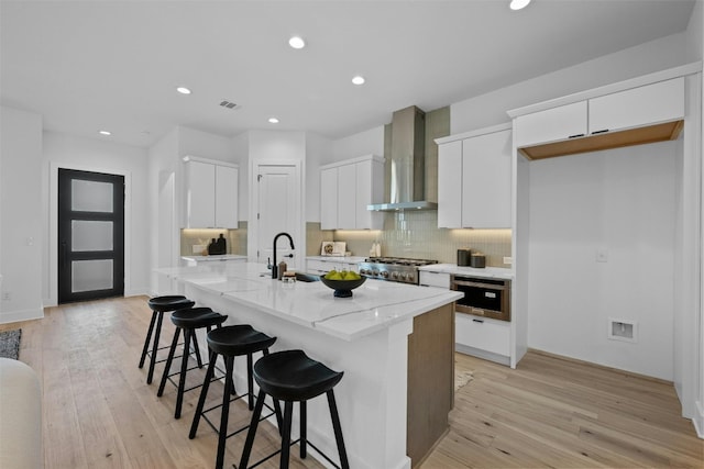 kitchen featuring light wood-type flooring, wall chimney exhaust hood, a breakfast bar, a kitchen island with sink, and white cabinets