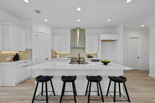 kitchen featuring a center island with sink, white cabinets, wall chimney range hood, and light hardwood / wood-style flooring