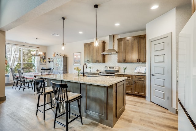 kitchen featuring sink, stainless steel range with electric stovetop, wall chimney range hood, light stone counters, and a center island with sink