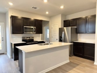 kitchen featuring sink, stainless steel appliances, dark brown cabinets, a center island with sink, and light wood-type flooring