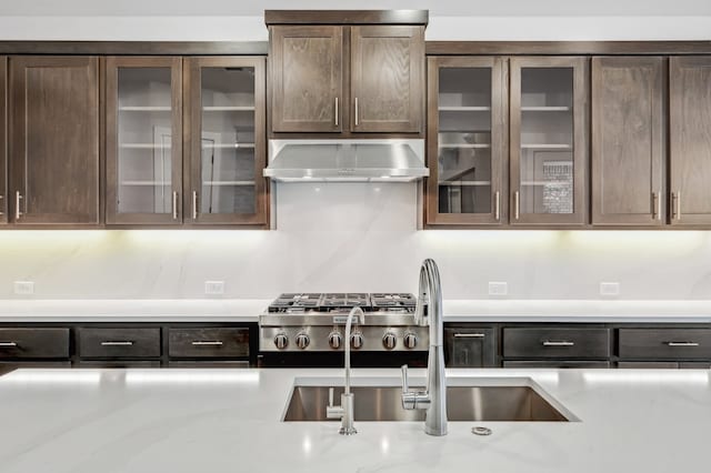 kitchen featuring dark brown cabinetry, backsplash, a sink, glass insert cabinets, and wall chimney exhaust hood