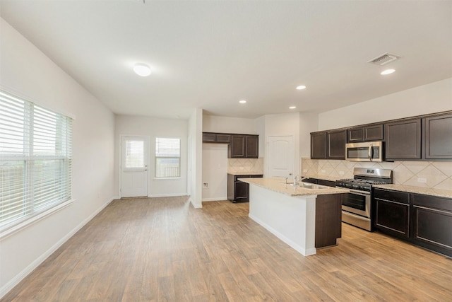 kitchen with light stone countertops, appliances with stainless steel finishes, light wood-type flooring, and a center island with sink