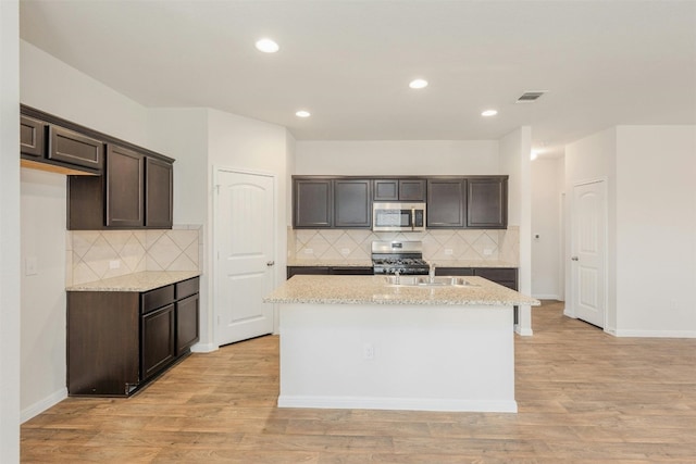 kitchen with dark brown cabinetry, light stone countertops, an island with sink, and stainless steel appliances