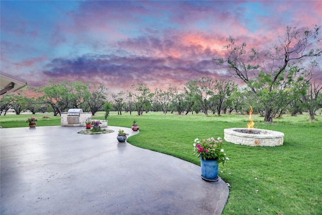 patio terrace at dusk featuring a fire pit, an outdoor kitchen, a grill, and a lawn