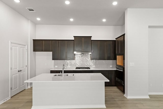 kitchen featuring black appliances, decorative backsplash, sink, light hardwood / wood-style flooring, and an island with sink