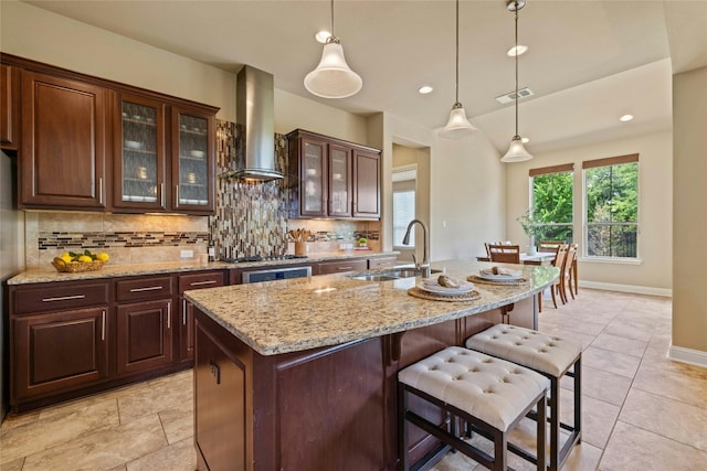 kitchen with sink, an island with sink, tasteful backsplash, and wall chimney range hood