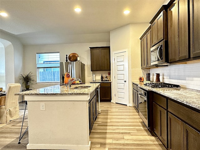 kitchen featuring appliances with stainless steel finishes, light stone counters, a kitchen island with sink, sink, and light hardwood / wood-style flooring