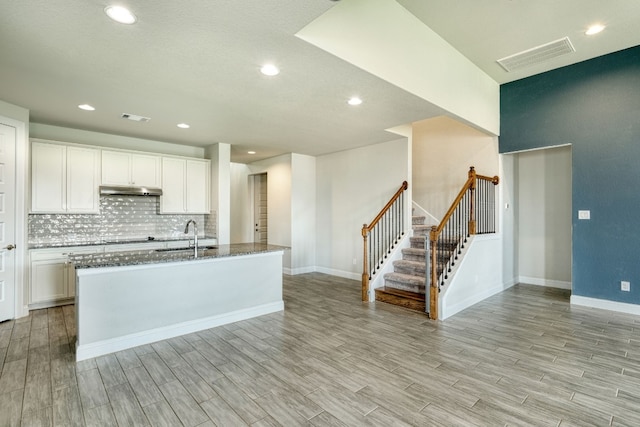 kitchen featuring a kitchen island with sink, sink, light hardwood / wood-style flooring, dark stone countertops, and white cabinets