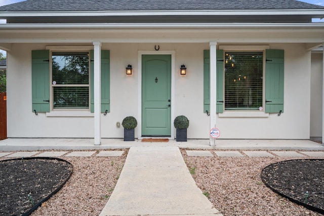 view of exterior entry featuring roof with shingles, covered porch, and stucco siding