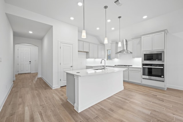 kitchen featuring sink, stainless steel appliances, wall chimney range hood, decorative light fixtures, and a kitchen island with sink