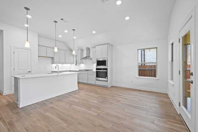 kitchen featuring a center island with sink, wall chimney range hood, hanging light fixtures, light hardwood / wood-style flooring, and appliances with stainless steel finishes