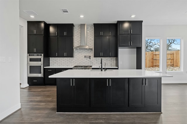 kitchen featuring double oven, sink, a kitchen island with sink, and wall chimney range hood