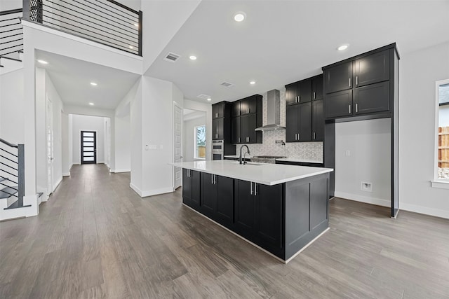 kitchen featuring wall chimney range hood, hardwood / wood-style flooring, sink, a kitchen island with sink, and oven