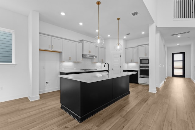 kitchen featuring stainless steel appliances, light hardwood / wood-style floors, white cabinetry, hanging light fixtures, and an island with sink