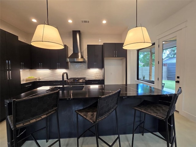 kitchen featuring decorative light fixtures, wall chimney range hood, a kitchen island with sink, and backsplash