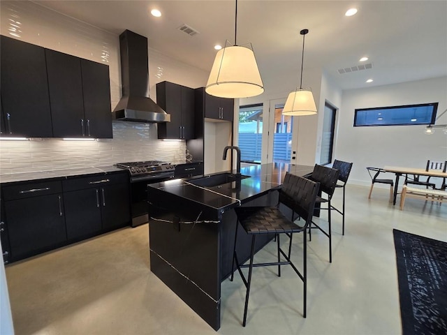 kitchen featuring decorative light fixtures, wall chimney range hood, backsplash, a center island with sink, and sink