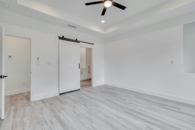 unfurnished bedroom featuring ceiling fan, a barn door, light hardwood / wood-style floors, and a tray ceiling