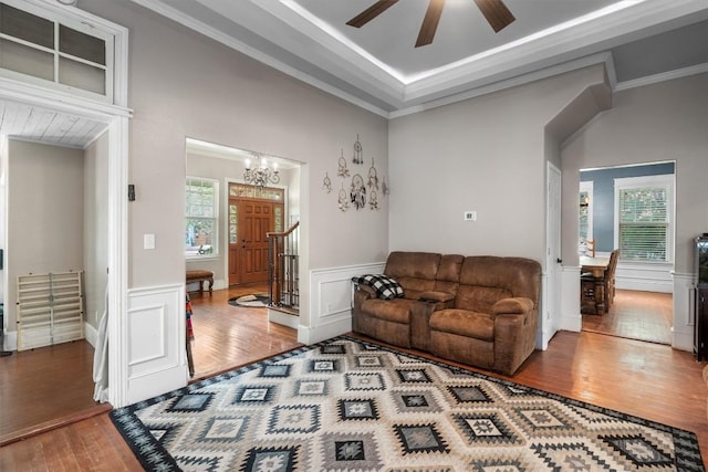 living room featuring wood-type flooring, ornamental molding, ceiling fan with notable chandelier, and a tray ceiling