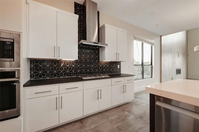 kitchen featuring white cabinets, appliances with stainless steel finishes, dark countertops, wall chimney range hood, and backsplash