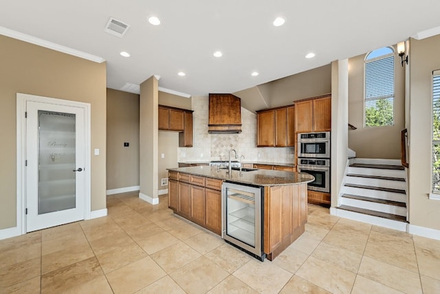 kitchen featuring beverage cooler, decorative backsplash, dark stone countertops, a center island with sink, and double oven