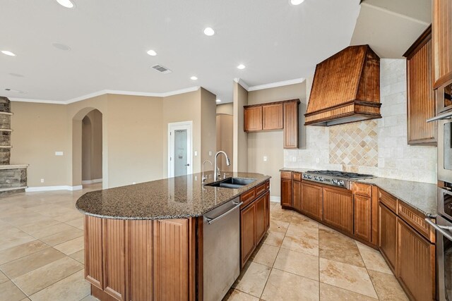 kitchen with appliances with stainless steel finishes, a center island with sink, sink, custom exhaust hood, and dark stone countertops