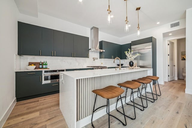 kitchen with a center island with sink, appliances with stainless steel finishes, hanging light fixtures, light countertops, and wall chimney range hood