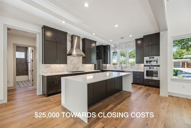 kitchen with a center island, wall chimney range hood, light hardwood / wood-style floors, decorative backsplash, and appliances with stainless steel finishes