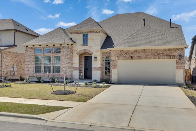 french country style house with driveway, a shingled roof, a garage, stone siding, and brick siding