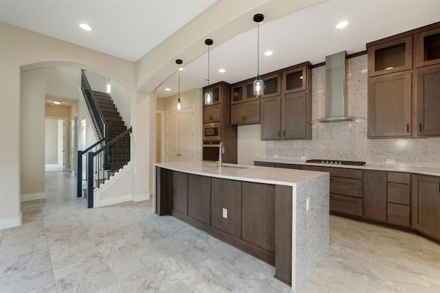 kitchen featuring decorative light fixtures, wall chimney range hood, stainless steel appliances, a center island with sink, and backsplash