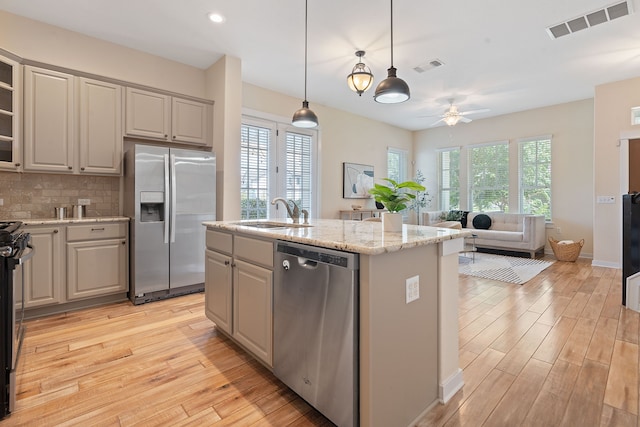 kitchen with tasteful backsplash, light wood-type flooring, appliances with stainless steel finishes, an island with sink, and light stone counters