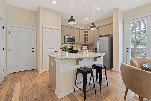 kitchen featuring appliances with stainless steel finishes, an island with sink, backsplash, hanging light fixtures, and light stone counters