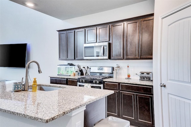 kitchen featuring sink, stainless steel appliances, dark brown cabinetry, light stone counters, and a center island with sink