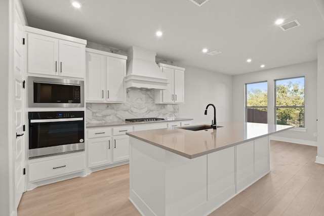 kitchen featuring appliances with stainless steel finishes, custom range hood, a kitchen island with sink, sink, and white cabinets