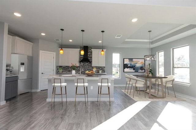 kitchen with white refrigerator with ice dispenser, a kitchen island with sink, hanging light fixtures, wall chimney exhaust hood, and a tray ceiling