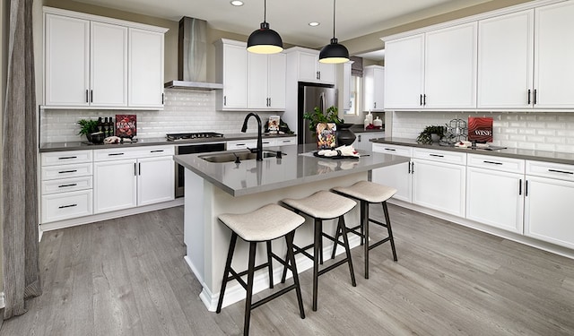 kitchen with light hardwood / wood-style floors, a center island with sink, hanging light fixtures, wall chimney range hood, and appliances with stainless steel finishes