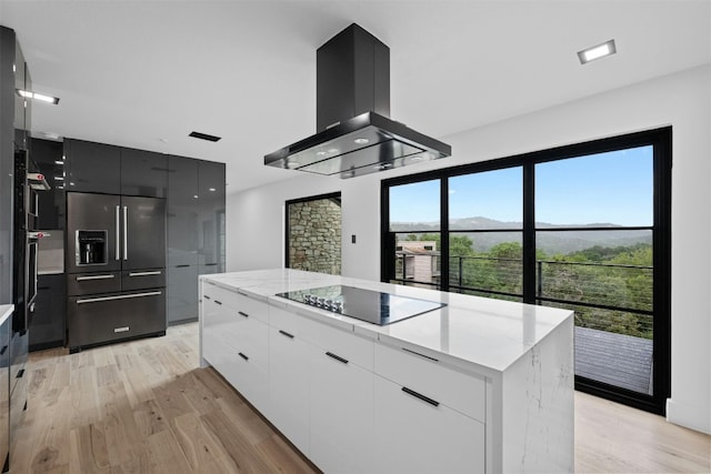 kitchen with black electric stovetop, island range hood, built in fridge, white cabinetry, and a wealth of natural light