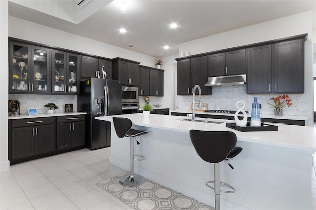 kitchen with a kitchen breakfast bar, a center island with sink, appliances with stainless steel finishes, light tile patterned floors, and backsplash