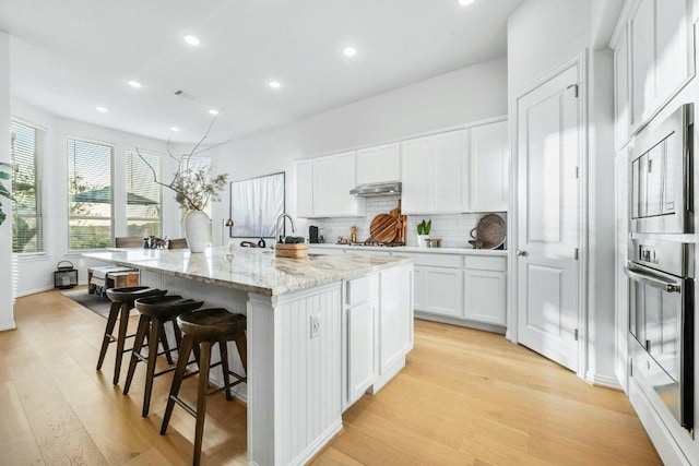 kitchen with light stone counters, a center island with sink, stainless steel appliances, white cabinets, and light hardwood / wood-style flooring