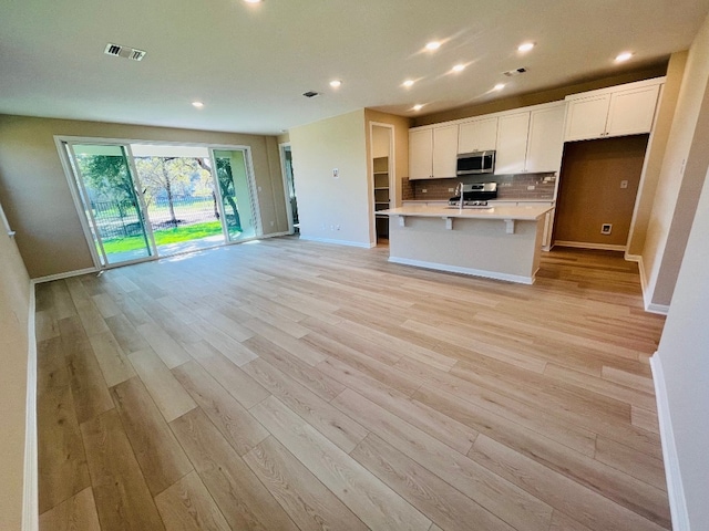 kitchen with stainless steel appliances, white cabinetry, a center island with sink, backsplash, and light hardwood / wood-style flooring