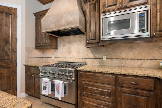 kitchen featuring premium range hood, decorative backsplash, light stone counters, stainless steel appliances, and dark brown cabinets