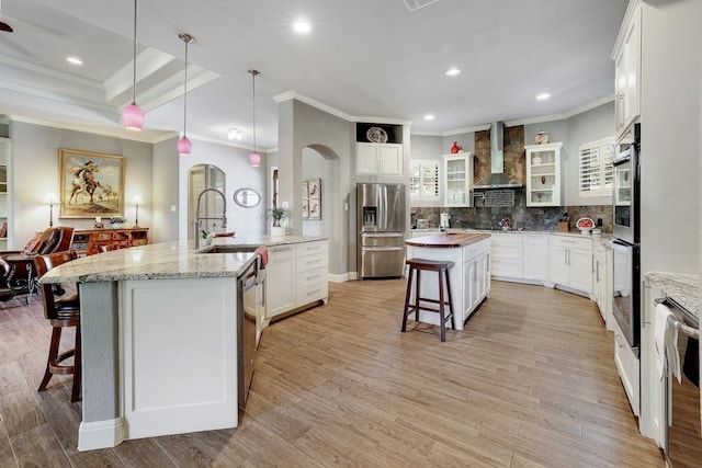 kitchen featuring stainless steel appliances, white cabinets, wall chimney exhaust hood, hanging light fixtures, and a center island with sink