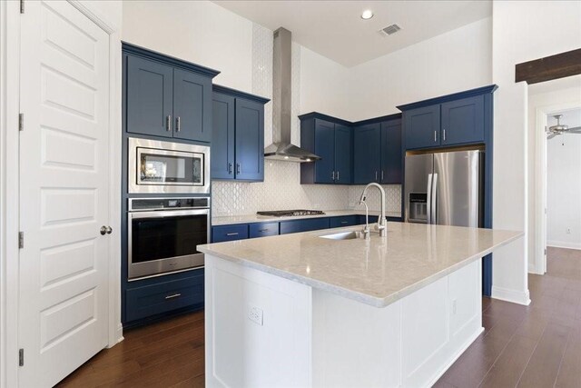 kitchen featuring blue cabinets, wall chimney exhaust hood, appliances with stainless steel finishes, sink, and a center island with sink