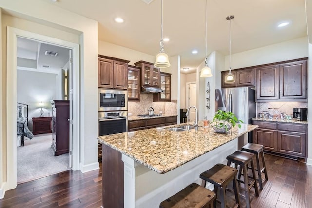 kitchen with a center island with sink, stainless steel appliances, hanging light fixtures, glass insert cabinets, and a sink