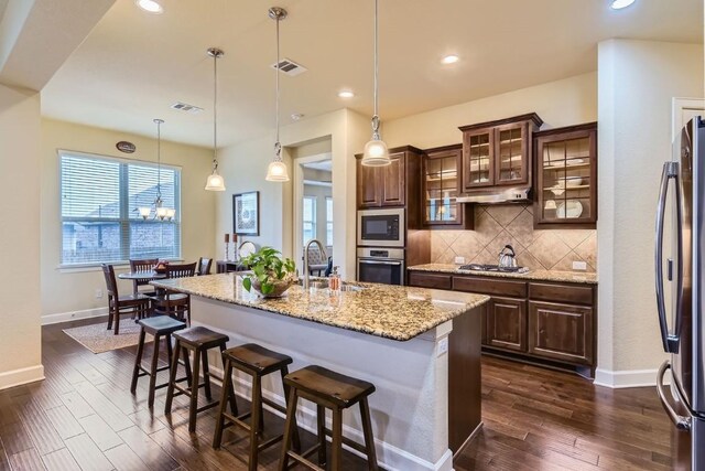 kitchen with an island with sink, stainless steel appliances, decorative backsplash, dark brown cabinets, and sink