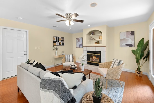 living room featuring a tile fireplace, built in shelves, ceiling fan, and hardwood / wood-style floors