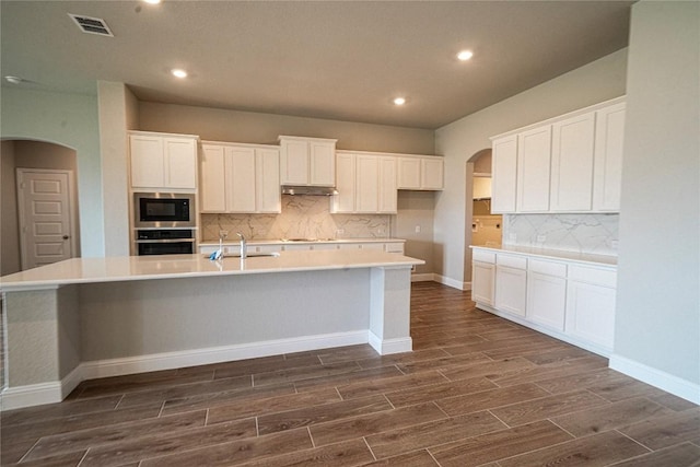 kitchen with wood finish floors, stainless steel oven, built in microwave, a large island with sink, and under cabinet range hood