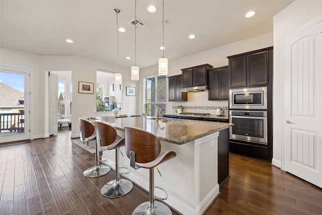 kitchen with stone counters, appliances with stainless steel finishes, pendant lighting, sink, and a kitchen island with sink