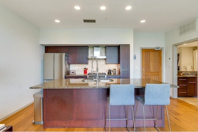 kitchen with light stone counters, stainless steel fridge, an island with sink, and wall chimney exhaust hood