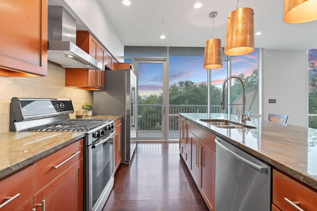 kitchen featuring light stone countertops, appliances with stainless steel finishes, wall chimney range hood, sink, and hanging light fixtures